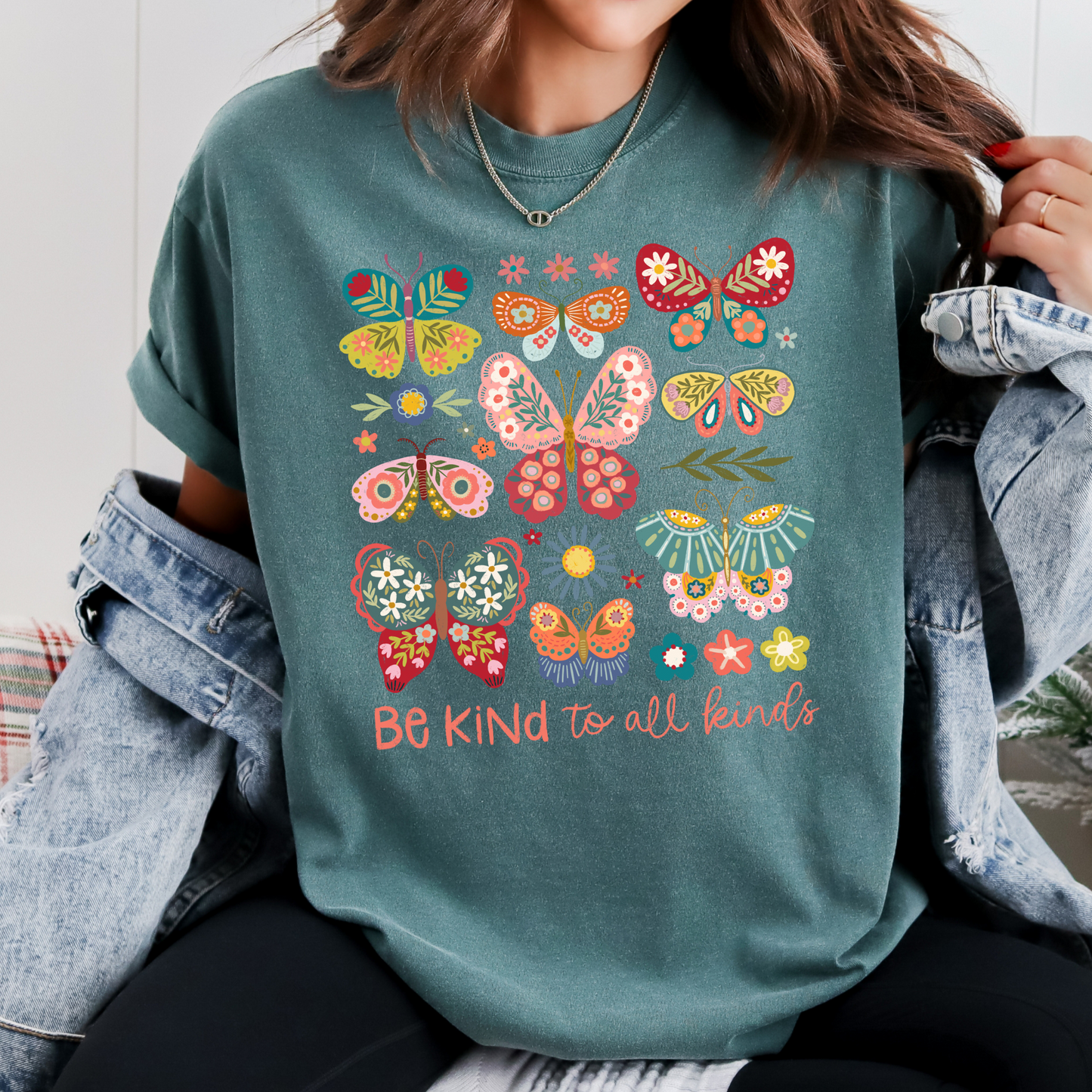 Person wearing a teal t-shirt with colorful butterfly design and 'Be Kind to all kinds' text, sitting in front of a Christmas tree.
