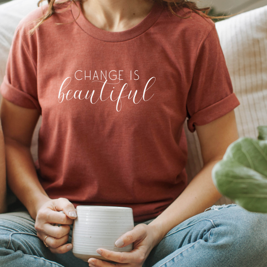 Person sitting on a couch holding a mug, wearing a t-shirt with 'Change is beautiful' text.