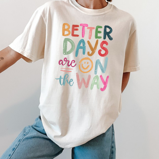 Person wearing a white t-shirt with colorful text with "Better Days Are On The Way" and blue jeans, standing against a plain wall.  